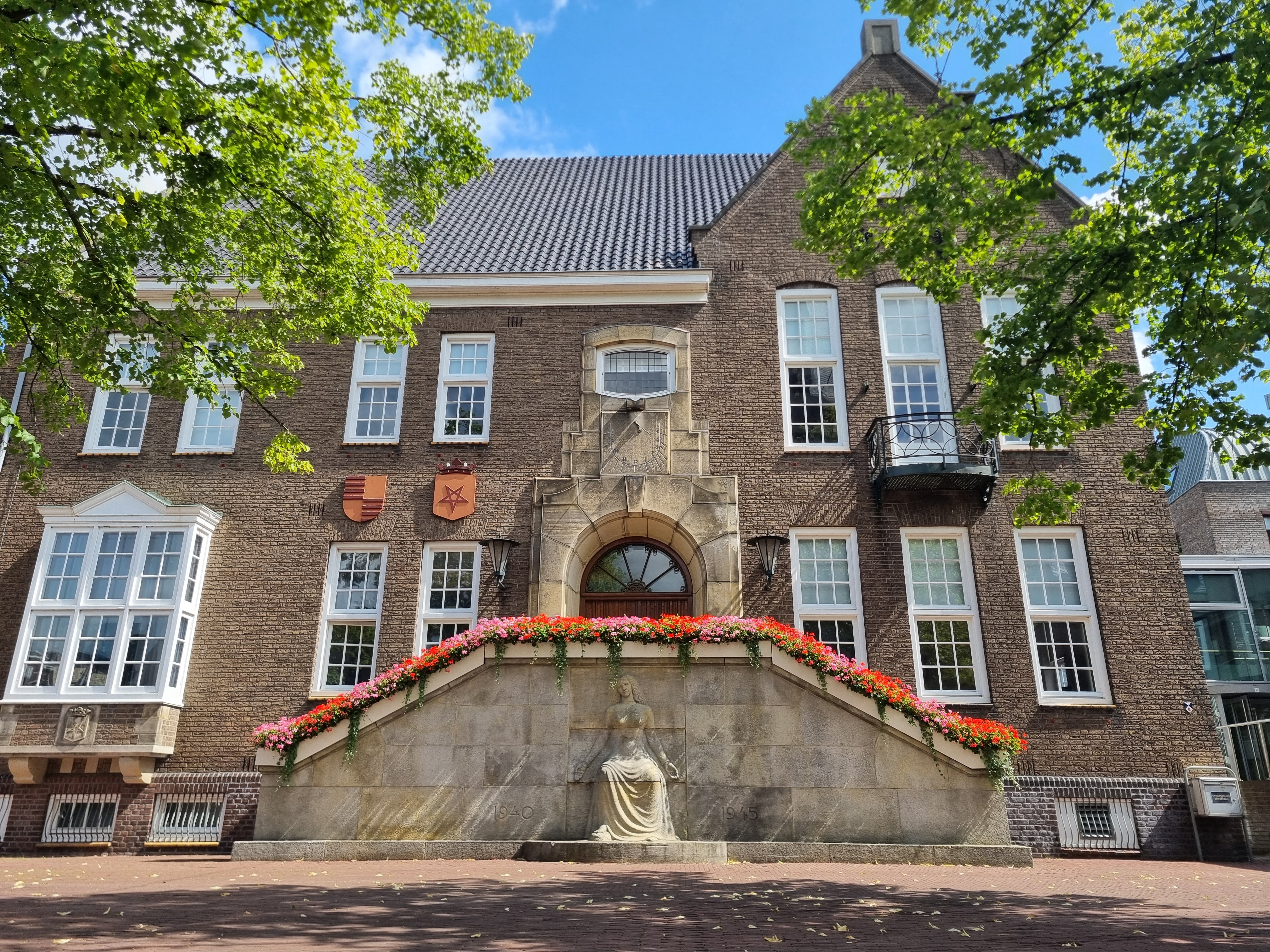 Voorkant oude gedeelte gemeentehuis vanaf marktplein gezien. Zonnig weer en bloemen op het bordes. 