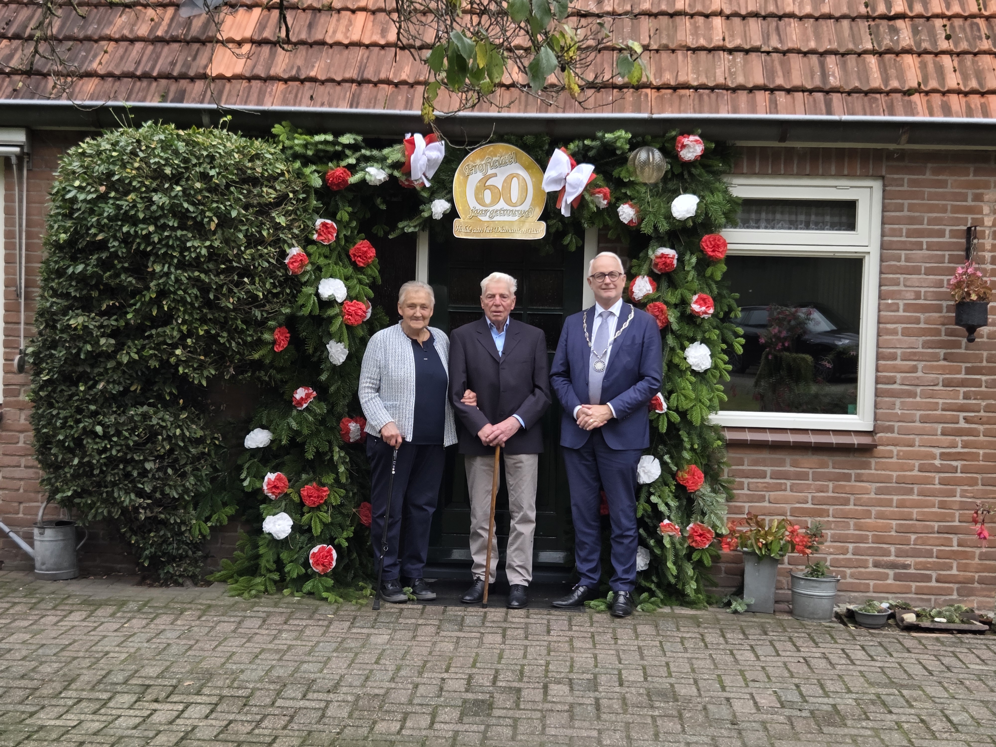 60-jarig bruidspaar Vedders poseert samen met burgemeester Gerard van den Hengel onder jubileumboog. 