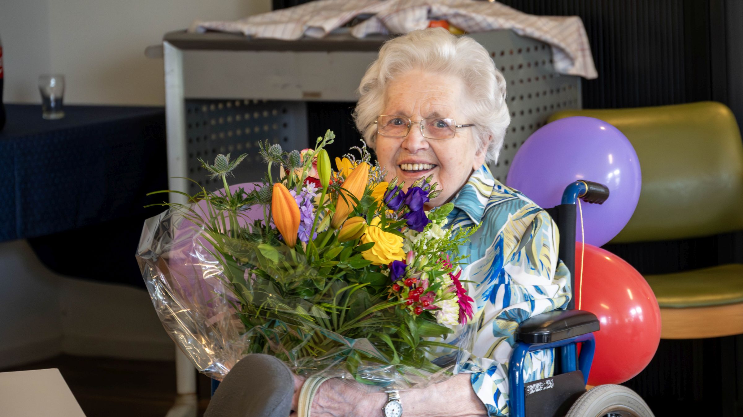 100-jarige Hermien ter Huurne-ten Asbroek met bos bloemen in met ballonnen versierde rolstoel. 