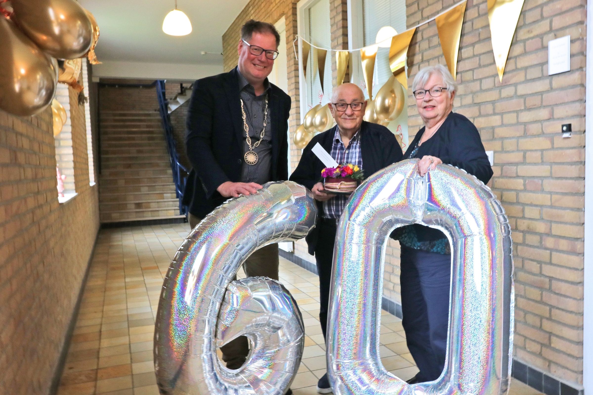 Wethouder Jan-Herman Scholten naast 60-jarig bruidspaar Serrano de la Torre. Voor hen het getal zestig in grote ballonnen.