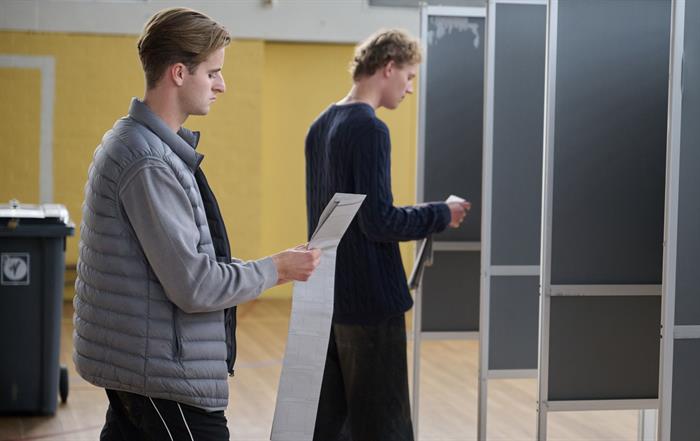 Twee jonge mannen met stembiljet op weg naar het stemhokje om te stemmen (stockfoto). 
