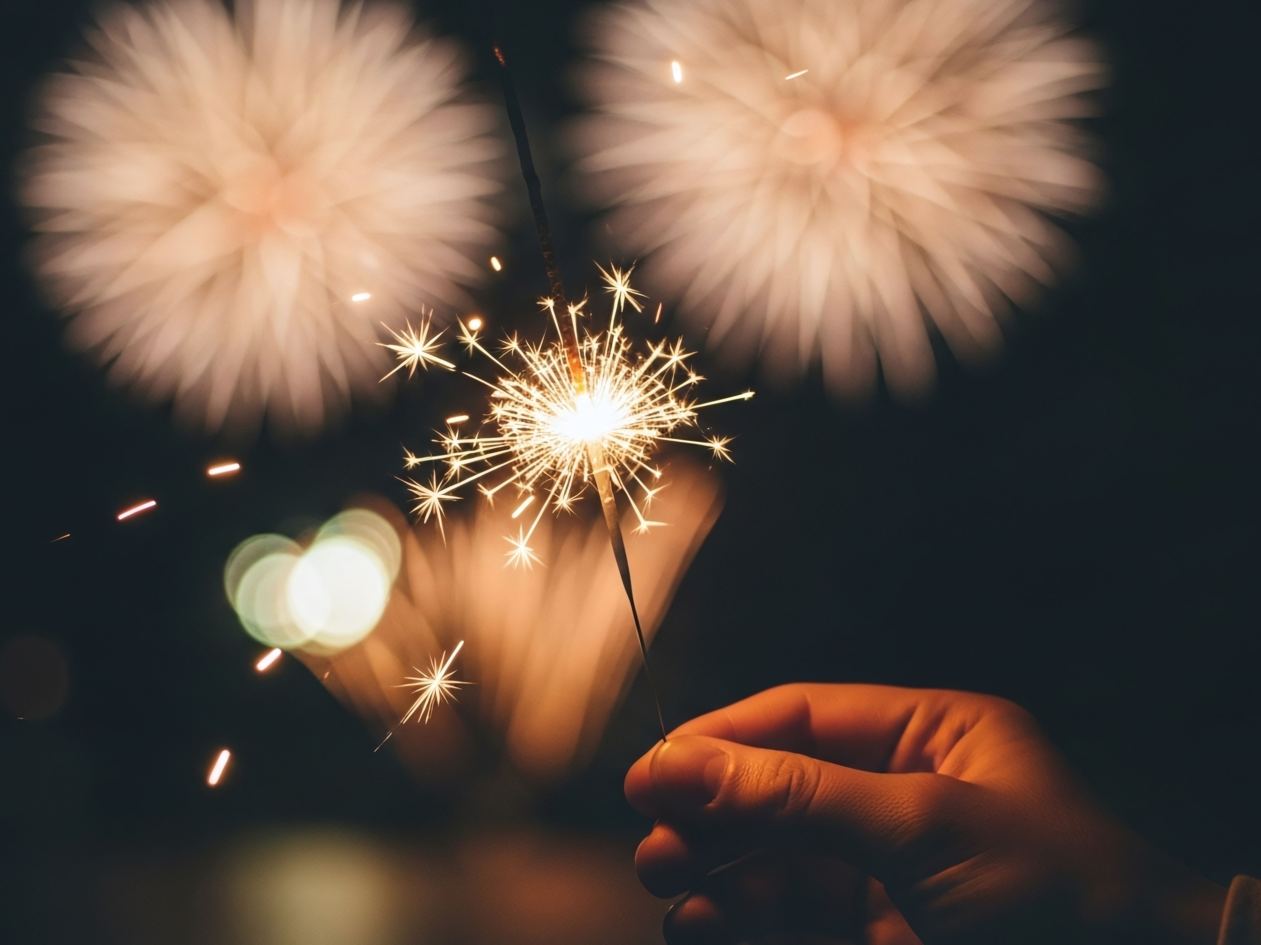 Foto van een hand die een brandend vuurwerksterretje vasthoudt. Op de achtergrond siervuurwerk zichtbaar in de lucht.