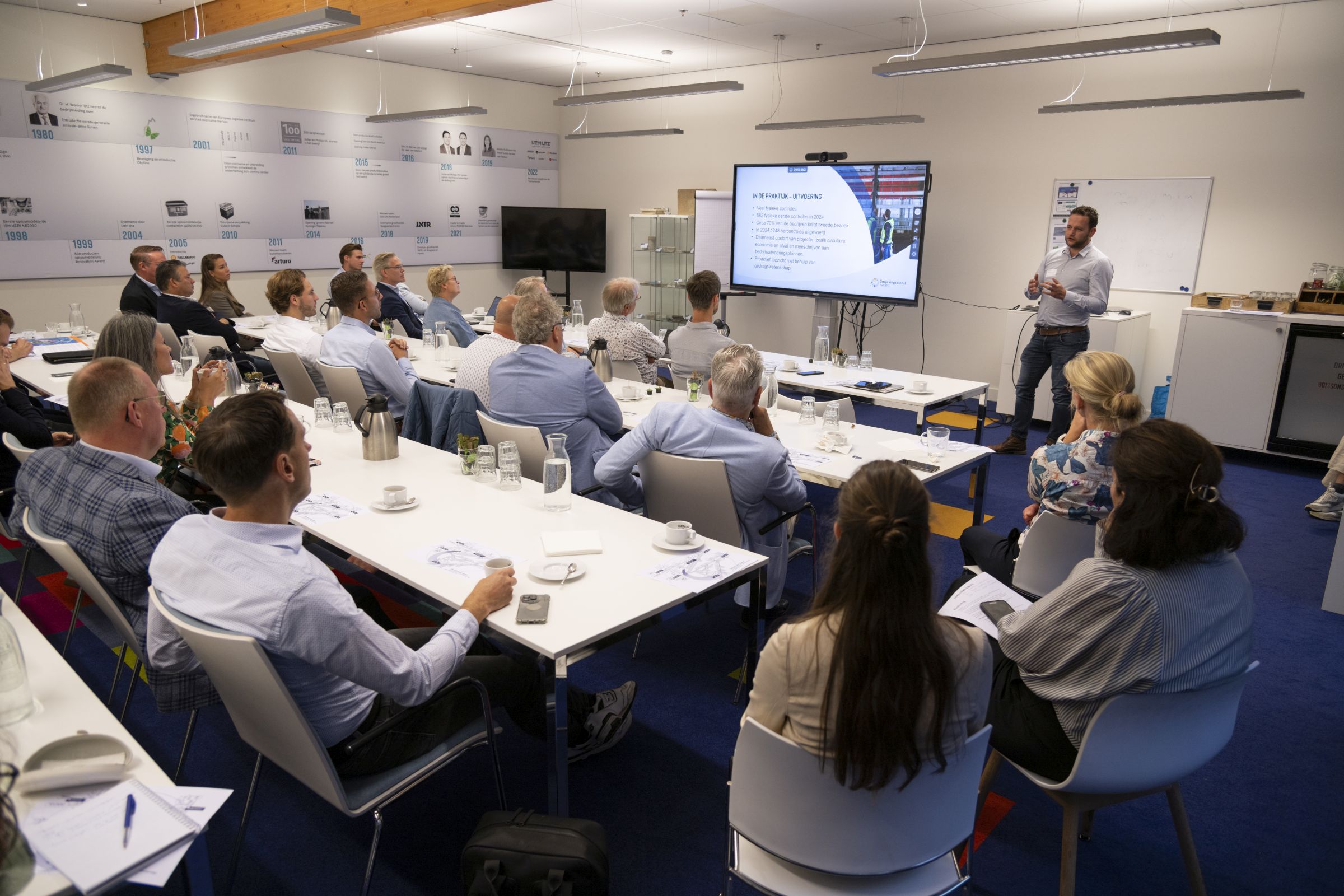 Groep mensen aan tafel luistert naar man die presentatie geeft. Hij staat naast een scherm. 