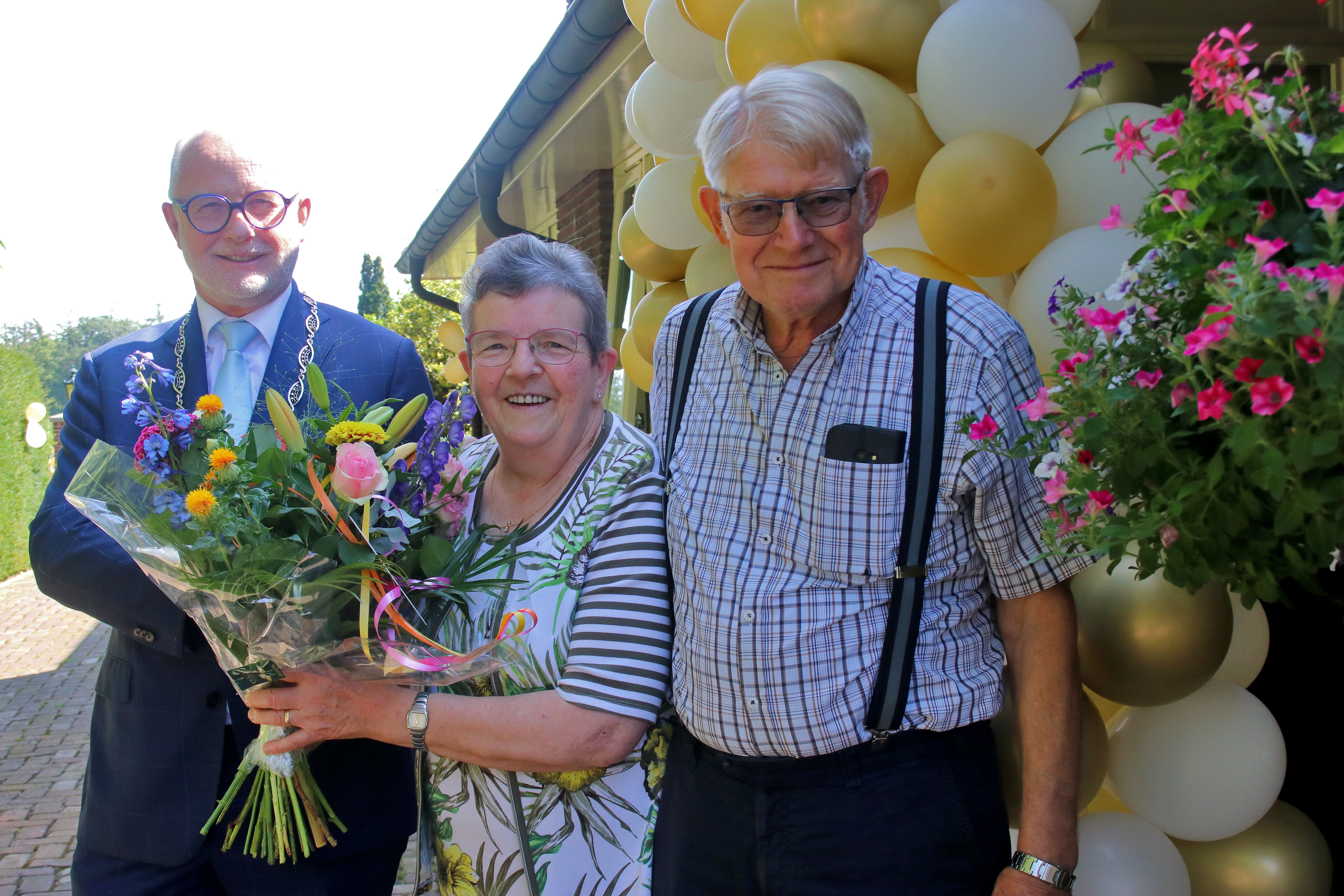 V.l.n.r. Burgemeester Gerard van den Hengel, Truus Veldhuis en Jan Breukers voor een boog met goude en witte ballonnen en een bos bloemen.