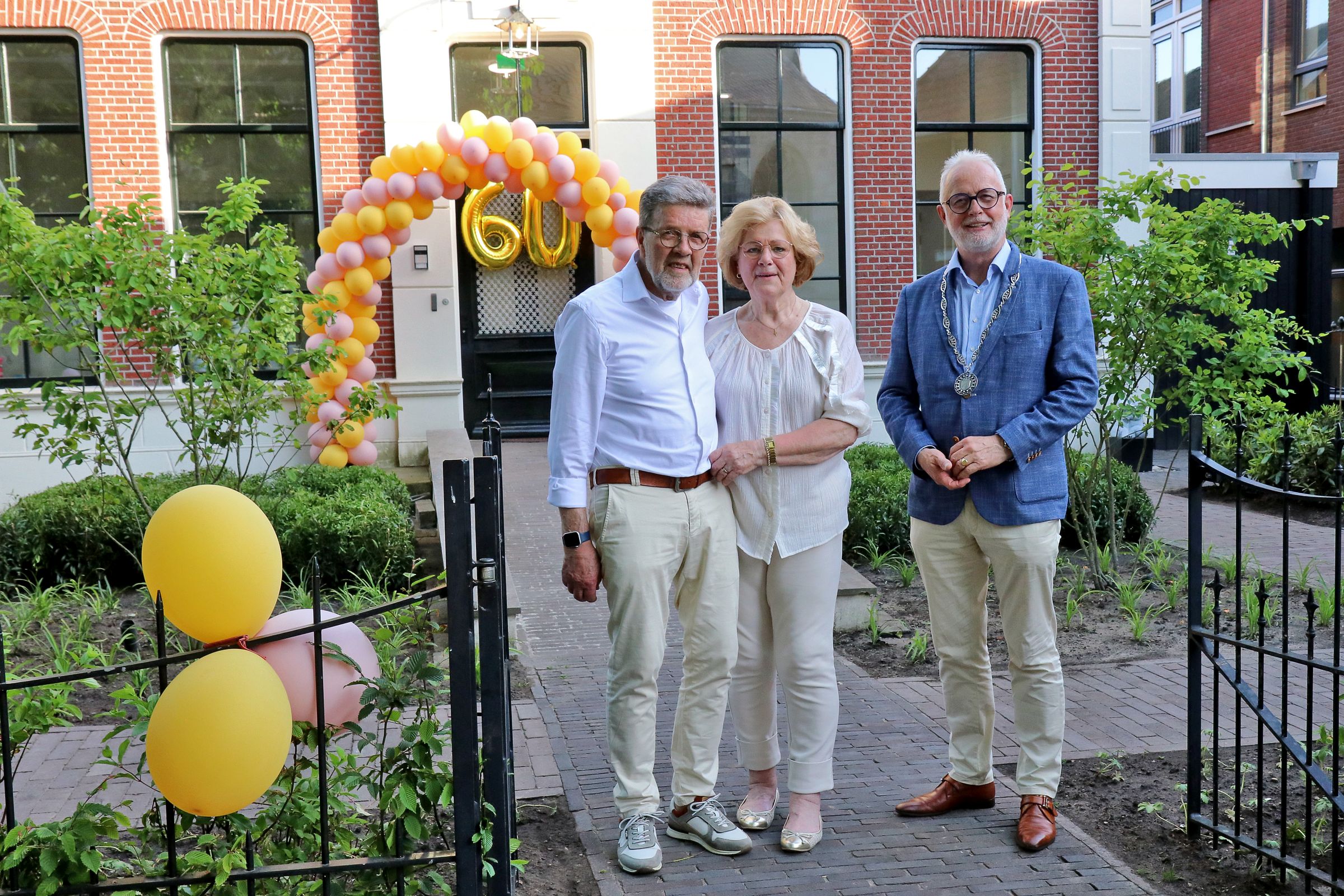 V.l.n.r. Rudi Keuper, Corrie Scheggetman en burgemeester Gerard van den Hengel voor de voornalige pastorie aan De Markt. Op de achtergrond een ballonnenboog en het getal 60 in ballonnen.  