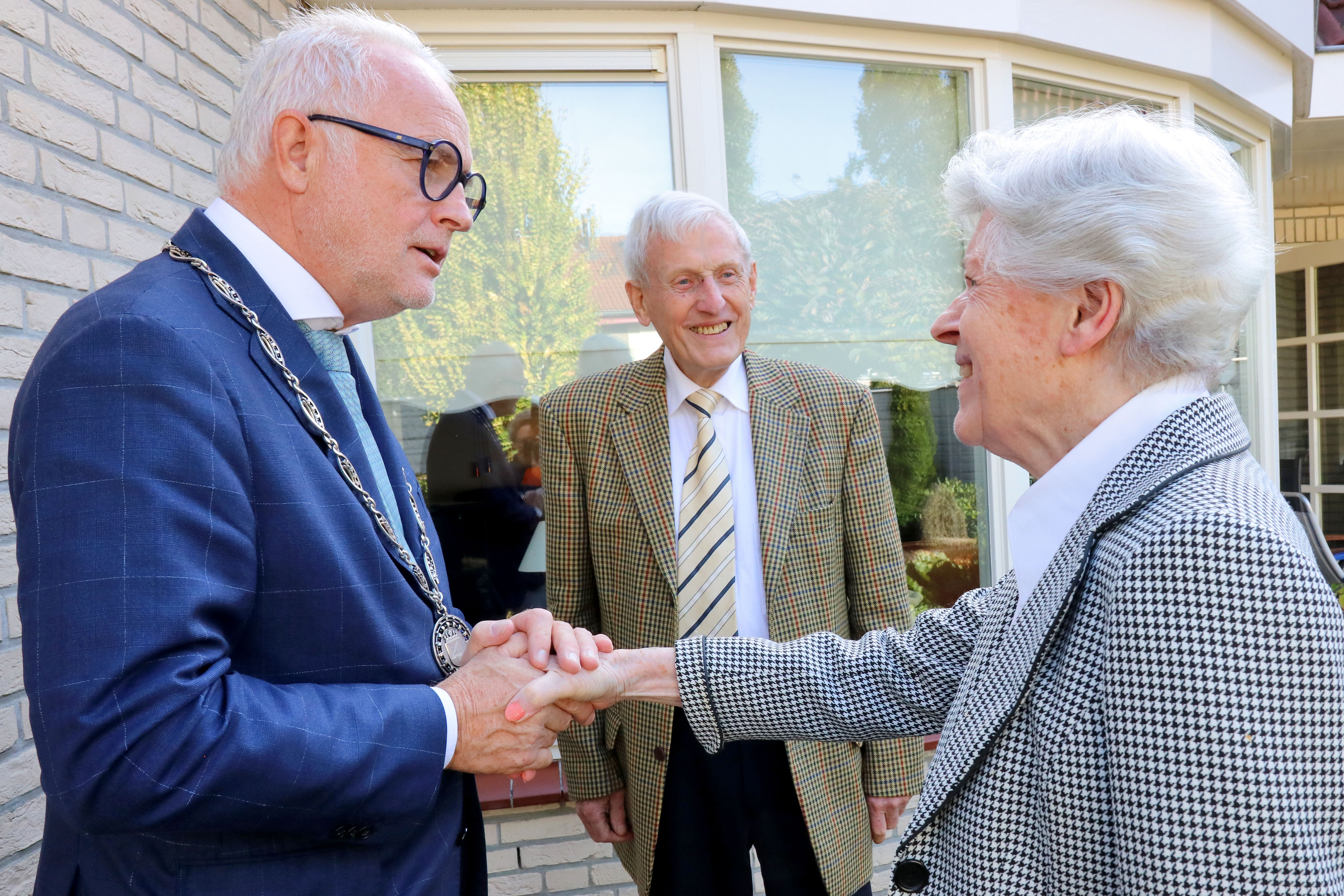 Burgemeester Gerard van den Hengel schudt de hand van Ardina Van Aken-Terpstra. Haar man Henk kijkt lachend toe.