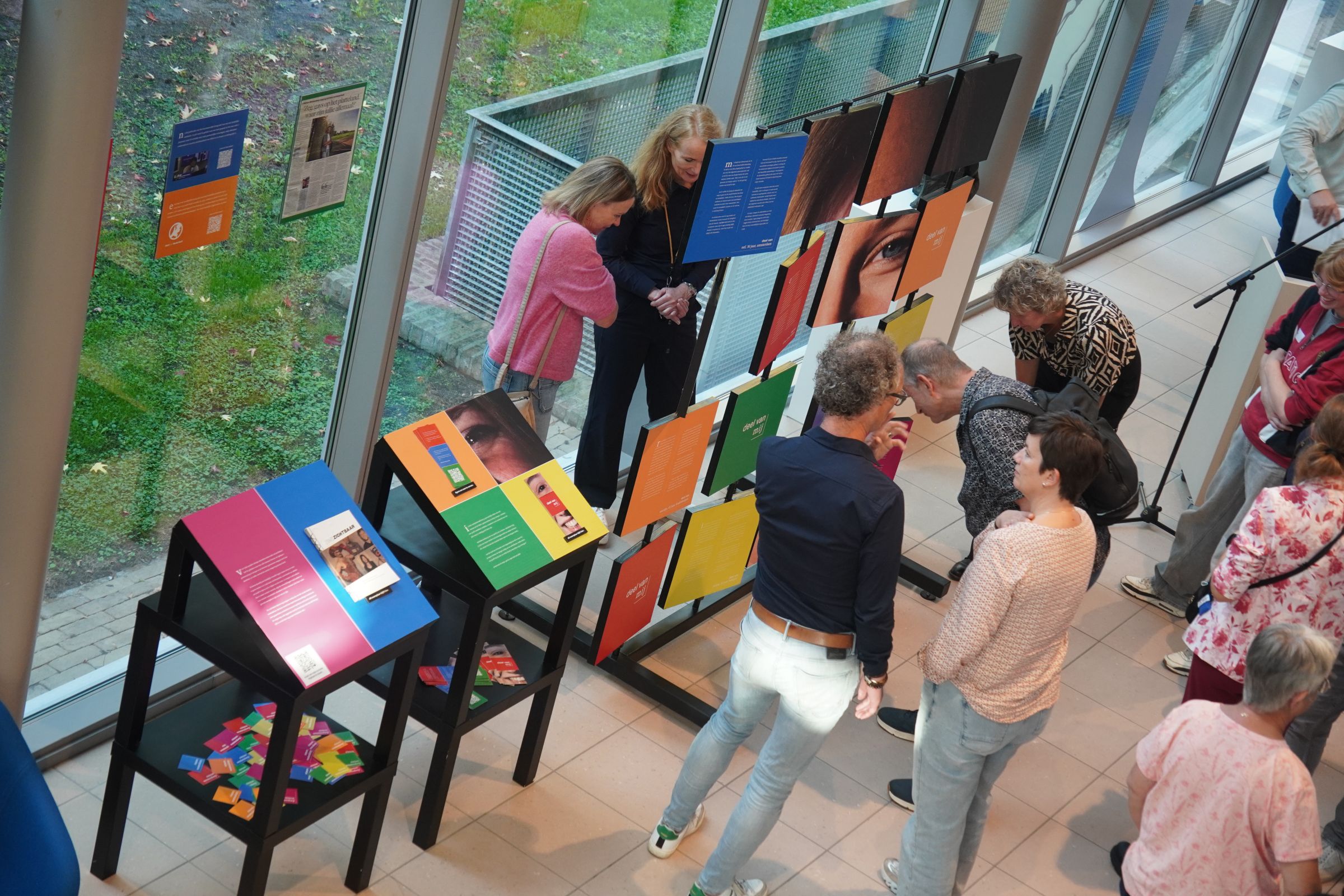 Mensen die binnen een tentoonstelling bekijken, met vierkante borden met daarop foto's, kleuren en verhalen.