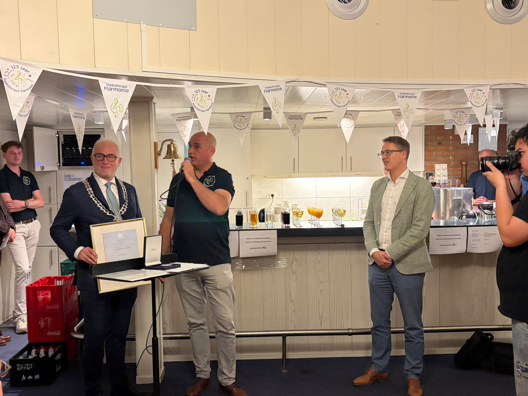 Burgemeester Gerard van den Hengel toont oorkonde tijden toespraak van lid Harmonie. Wethouder Thij Looman luistert mee. 