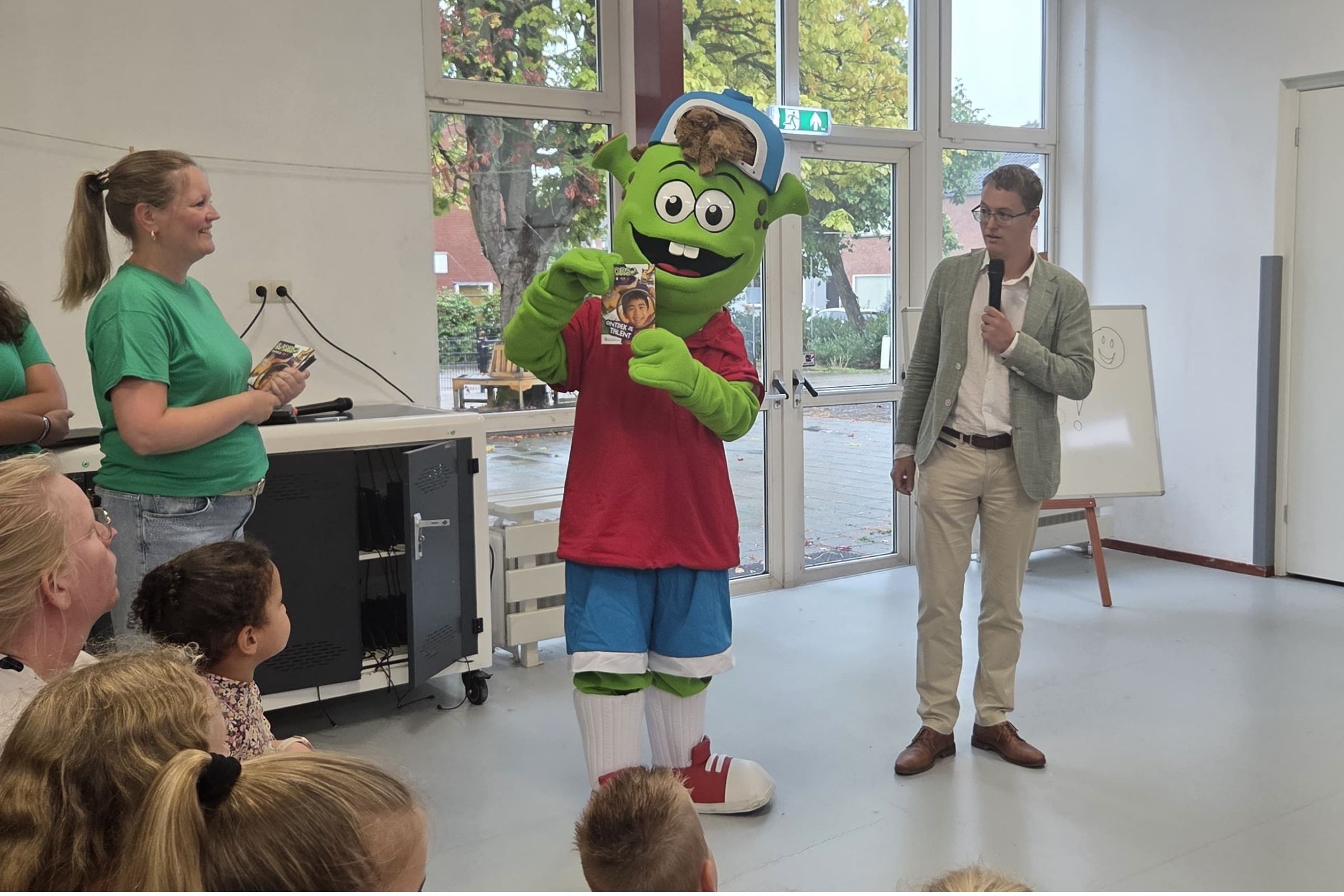 De Sjors mascotte deelt het eerste Sjors-boekje uit, met wethouder Thij Looman naast zich.